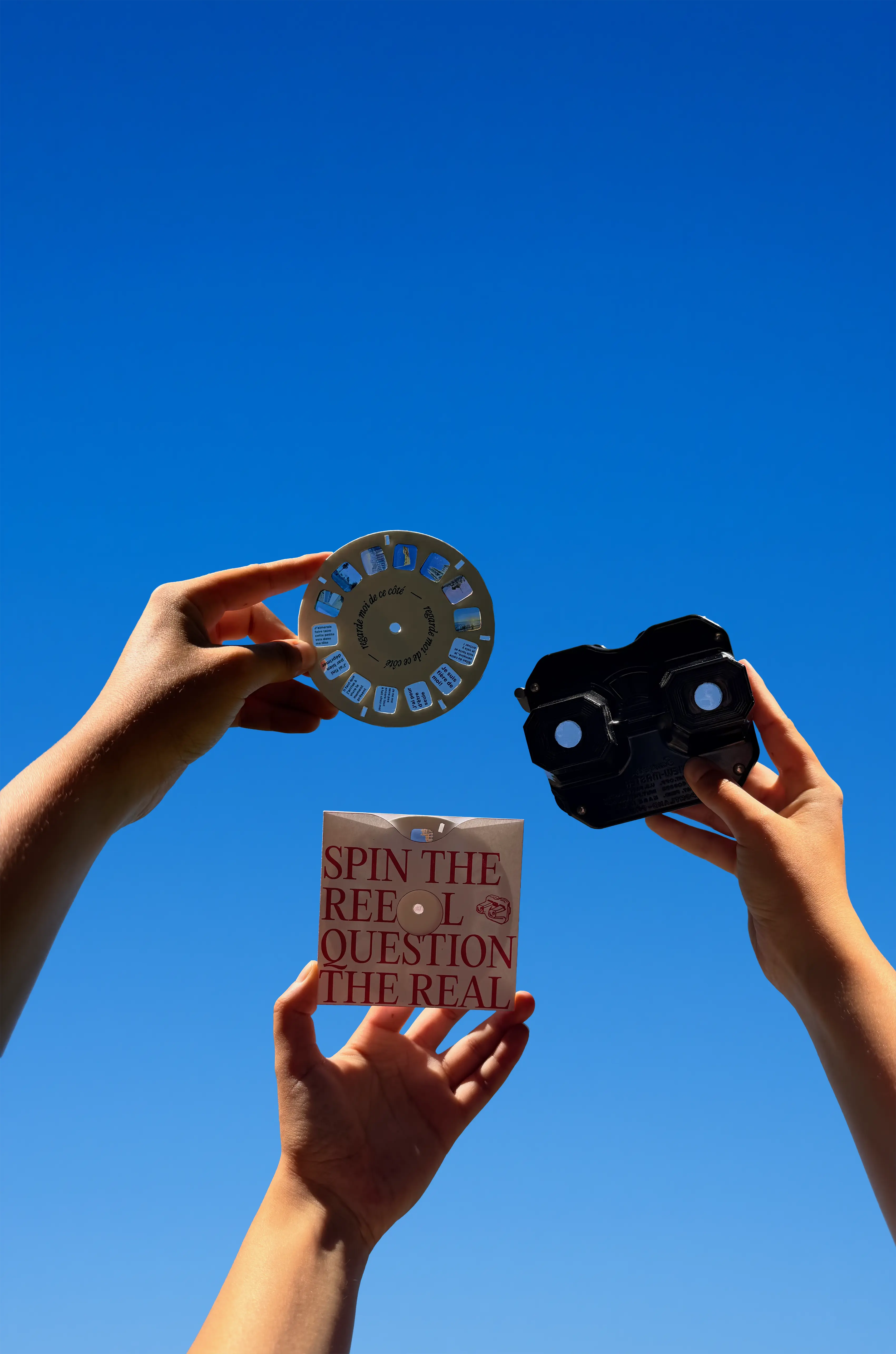 Deux mains tendues vers le ciel bleu tenant les bobines, le View-Master et une pochette